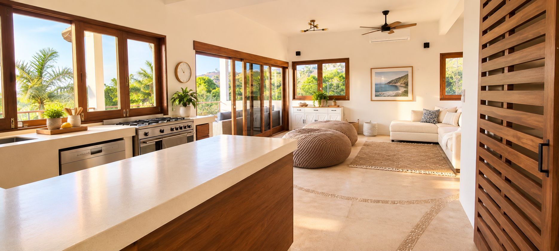 A modern kitchen with a large island, a dining area with a sofa and ottoman, and a view of palm trees through large windows.