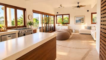 A modern kitchen with a large island, a dining area with a sofa and ottoman, and a view of palm trees through large windows.