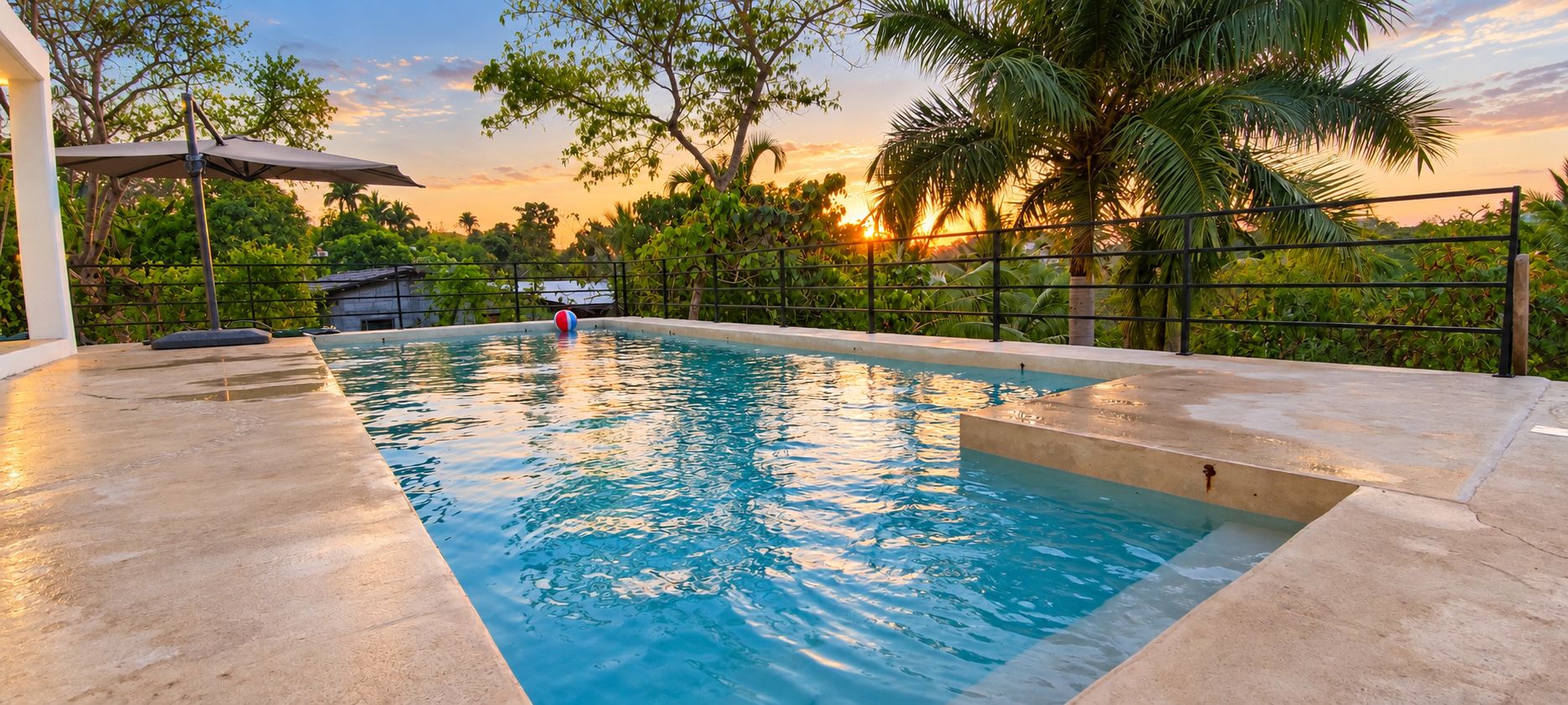 A swimming pool with a sunset view, surrounded by lush greenery and a fence.