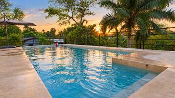 A swimming pool with a sunset view, surrounded by lush greenery and a fence.