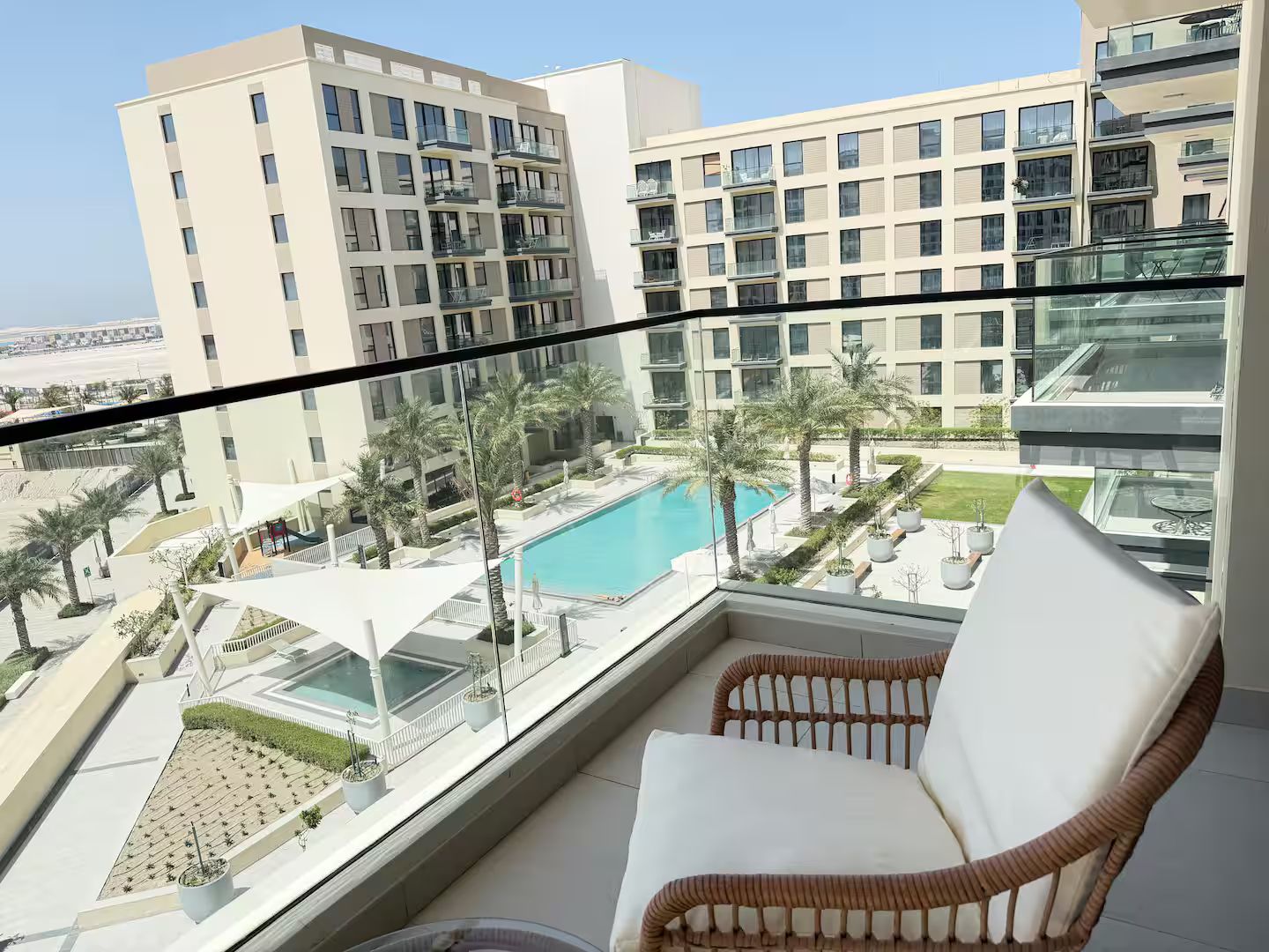 Un balcon avec un fauteuil en osier, donnant sur un espace piscine agrémenté de palmiers et sur un ensemble de bâtiments en arrière-plan.