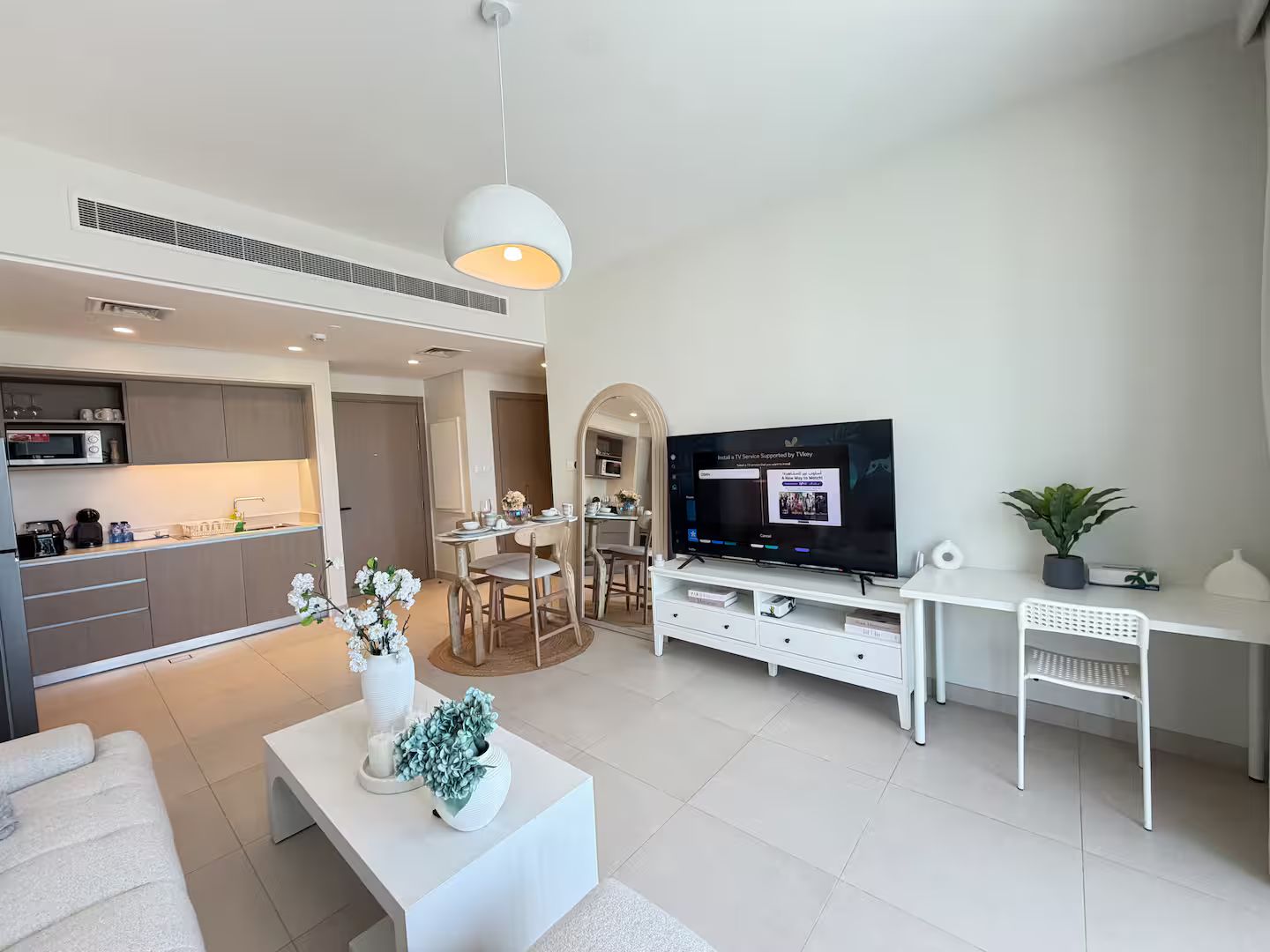 A modern living room with a flat-screen TV, white furniture, and a dining area with a kitchen in the background.