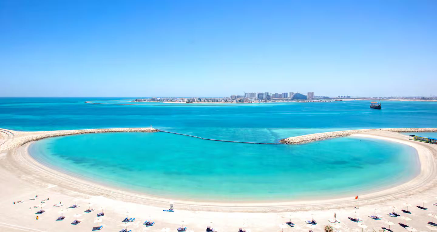A coastal beach with a clear blue sea, a sandy shore, and a distant cityscape.