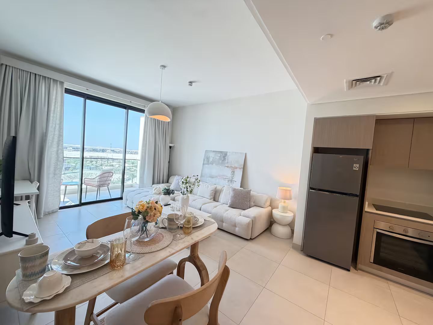 Una cocina moderna con horno y refrigerador integrados, un comedor con una mesa lista para dos comensales, y una sala de estar con sofá y un cuadro.