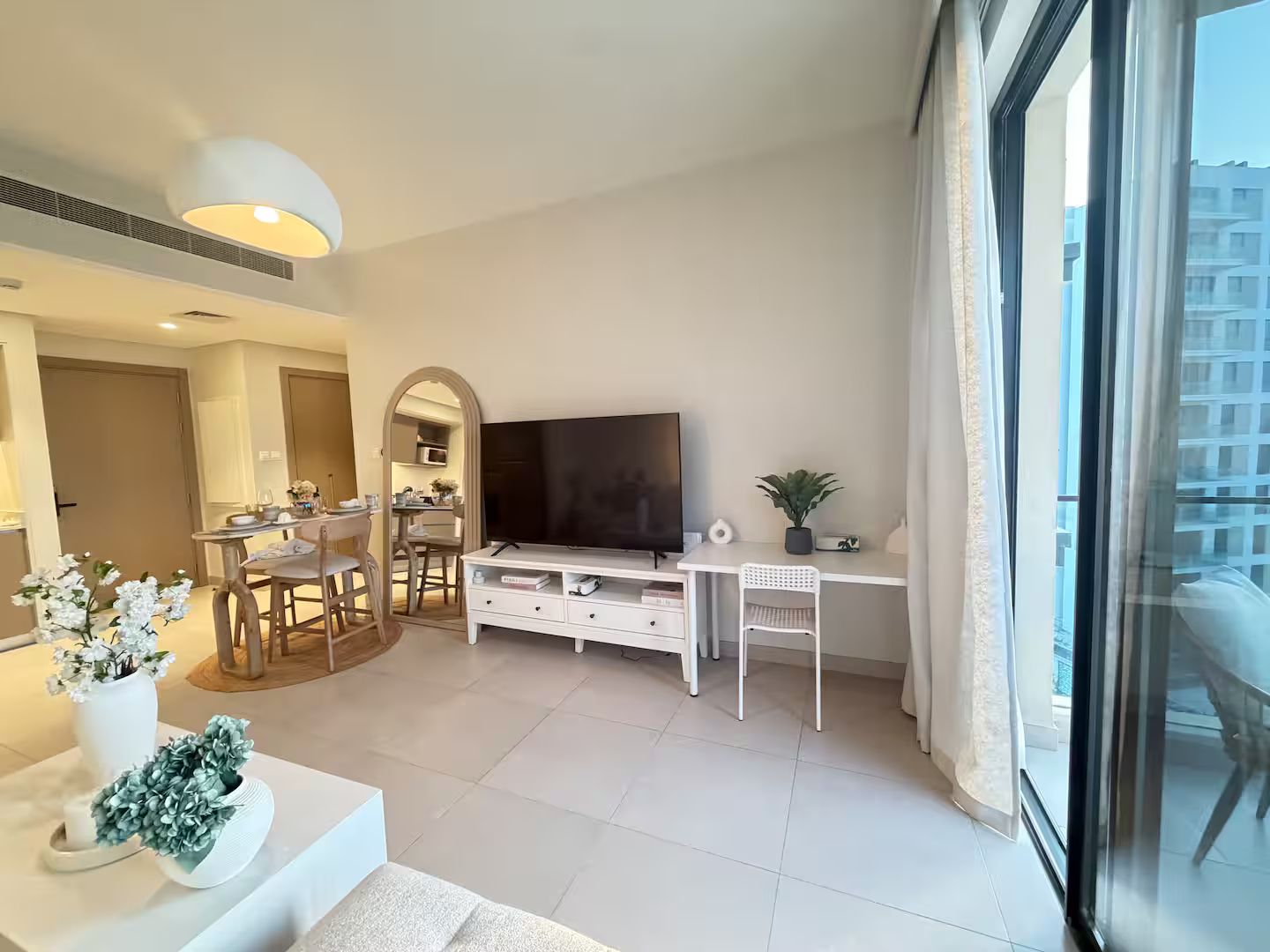 A modern living room with a flat-screen TV, white furniture, and a dining area visible through an archway.