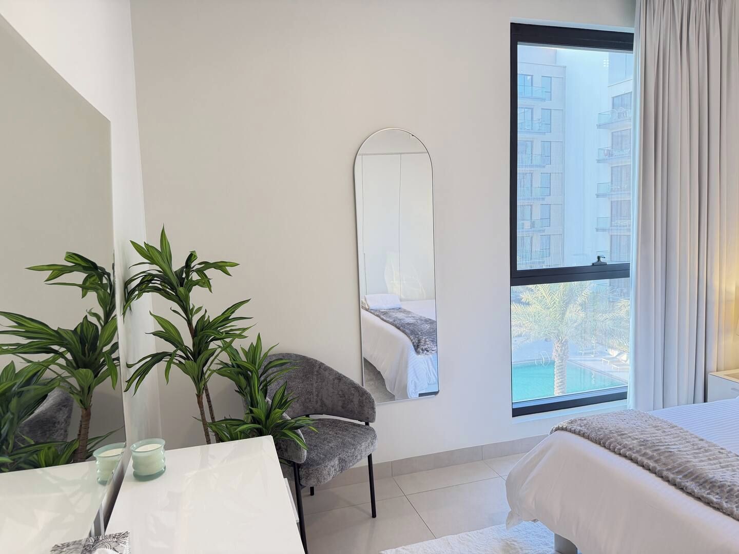 A hotel room with a bed, a chair, a table with a candle, a mirror, and a window with a view of buildings.