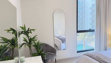 A hotel room with a bed, a chair, a table with a candle, a mirror, and a window with a view of buildings.