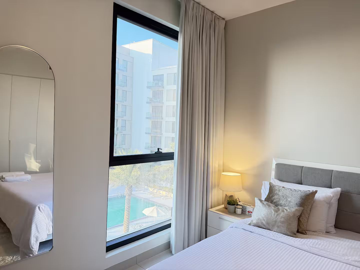 Une chambre d’hôtel avec un lit, une table de chevet avec une lampe, et une vue sur un immeuble et une piscine, le tout visible à travers une grande fenêtre.