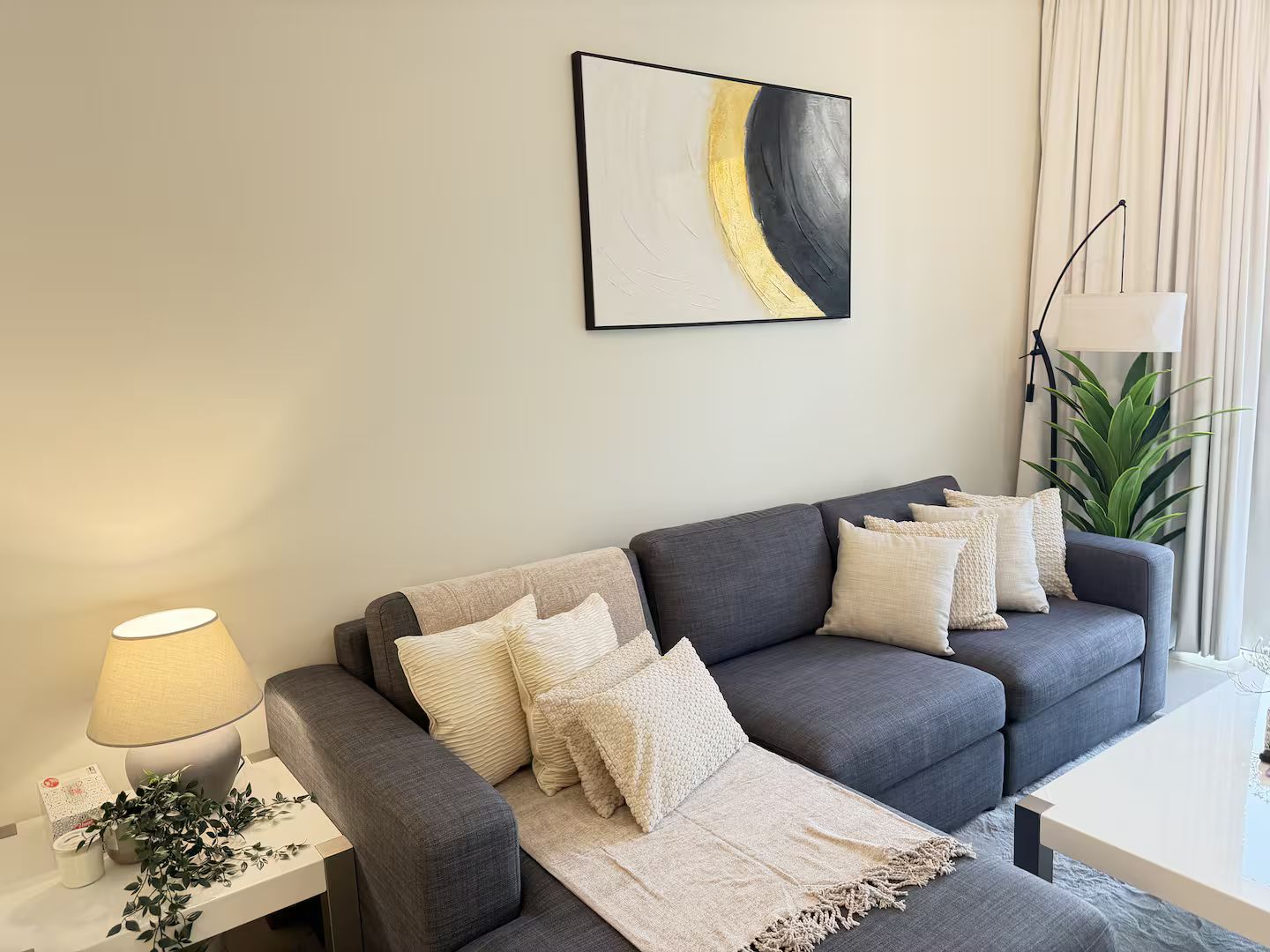 Ein modernes Wohnzimmer mit einem grauen Sofa, einem weißen Beistelltisch, einer Stehlampe und einem großen abstrakten Gemälde an der Wand.