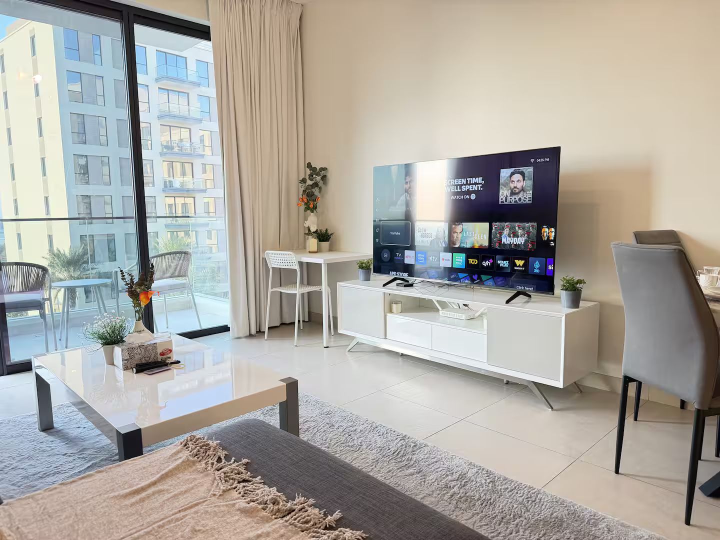 A modern living room with a large flat-screen TV, a white entertainment unit, a sofa, a coffee table, and a balcony with outdoor seating.
