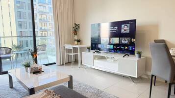 A modern living room with a large flat-screen TV, a white entertainment unit, a sofa, a coffee table, and a balcony with outdoor seating.