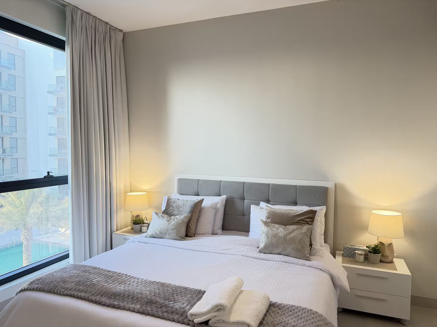 A hotel room with a bed, two bedside tables, a window with curtains, and a view of buildings outside.
