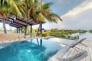 Ein Pool ohne Ende mit einer Holzterrasse, Palmen und Blick auf ruhiges Wasser.