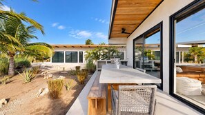 Un pati exterior modern amb una taula de fusta i cadires de vimet, envoltat d'un jardí ben cuidat amb diverses plantes i una palmera.