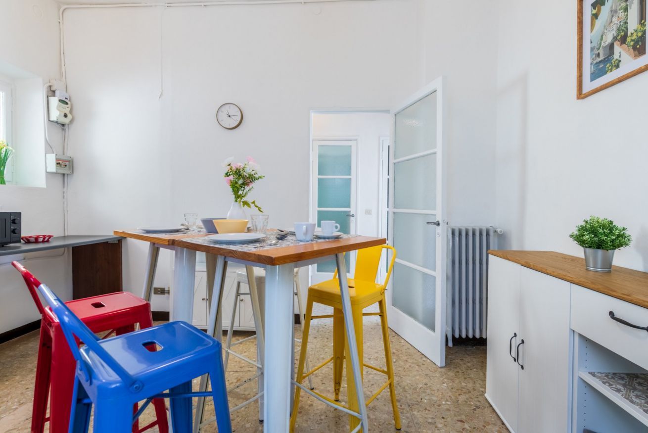 Eine kleine Küchenzeile mit einer weißen Arbeitsplatte, einem roten und blauen Stuhl, einem gelben Hocker, einem weißen Schrank und einer Uhr an der Wand.