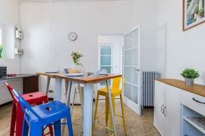Eine kleine Küchenzeile mit einer weißen Arbeitsplatte, einem roten und blauen Stuhl, einem gelben Hocker, einem weißen Schrank und einer Uhr an der Wand.