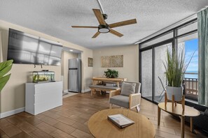 A modern living room with a ceiling fan, a dining area with a table and chairs, a kitchen with a refrigerator, and a large window offering a view.