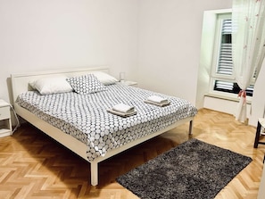 A neatly made bed with a patterned bedspread, a bedside table, and a window with blinds. - Luxury 100 m2, 2-bedroom apartment with balcony & garden (Rijeka)