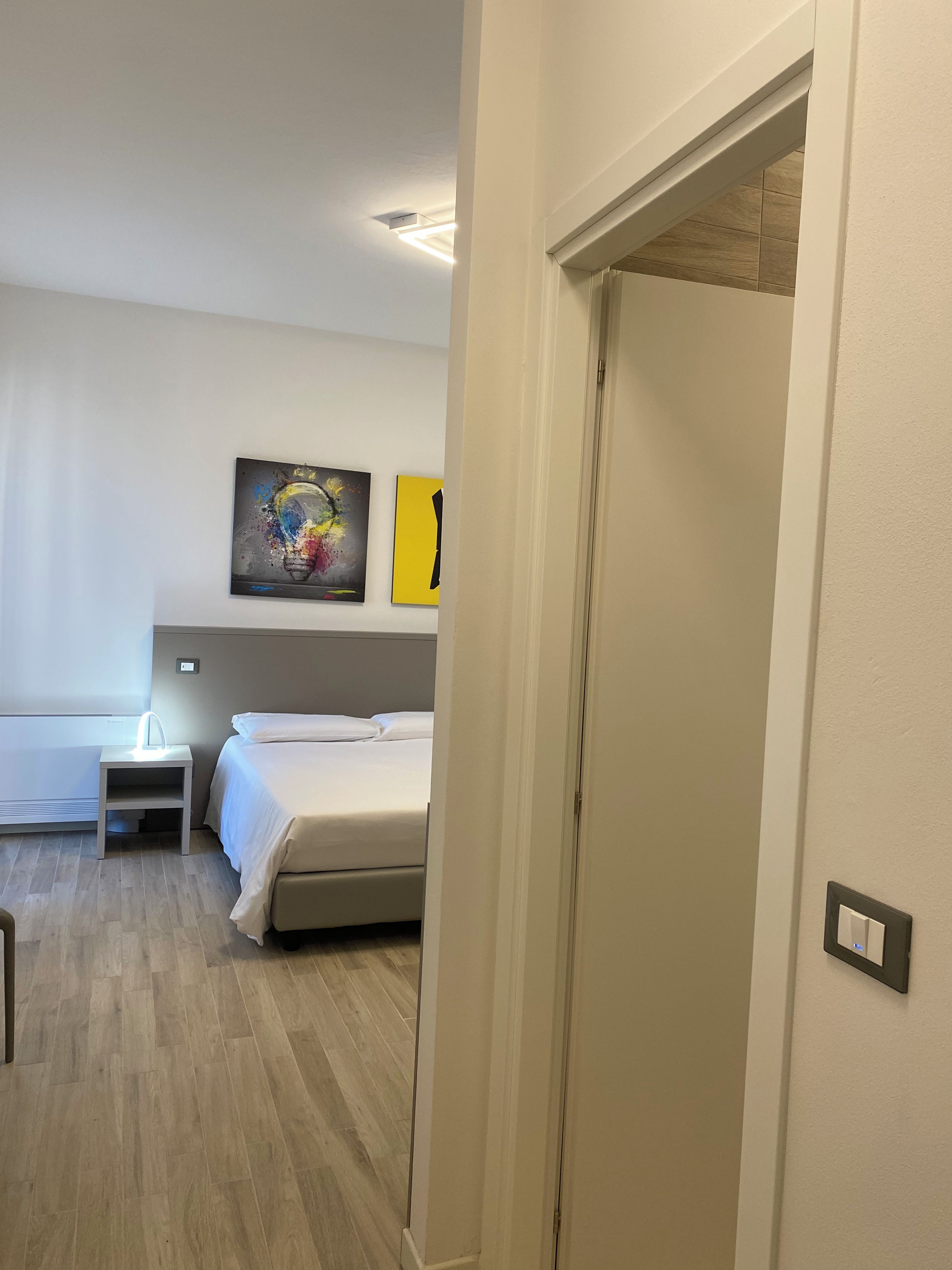 A hotel room with a bed, bedside table, and a small stool. There are two paintings on the wall and a ceiling light.