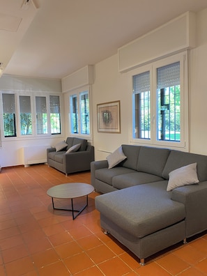 A spacious living room with a grey sectional sofa, a round coffee table, and large windows with blinds. - Villanova 4 rooms  (Villanova)