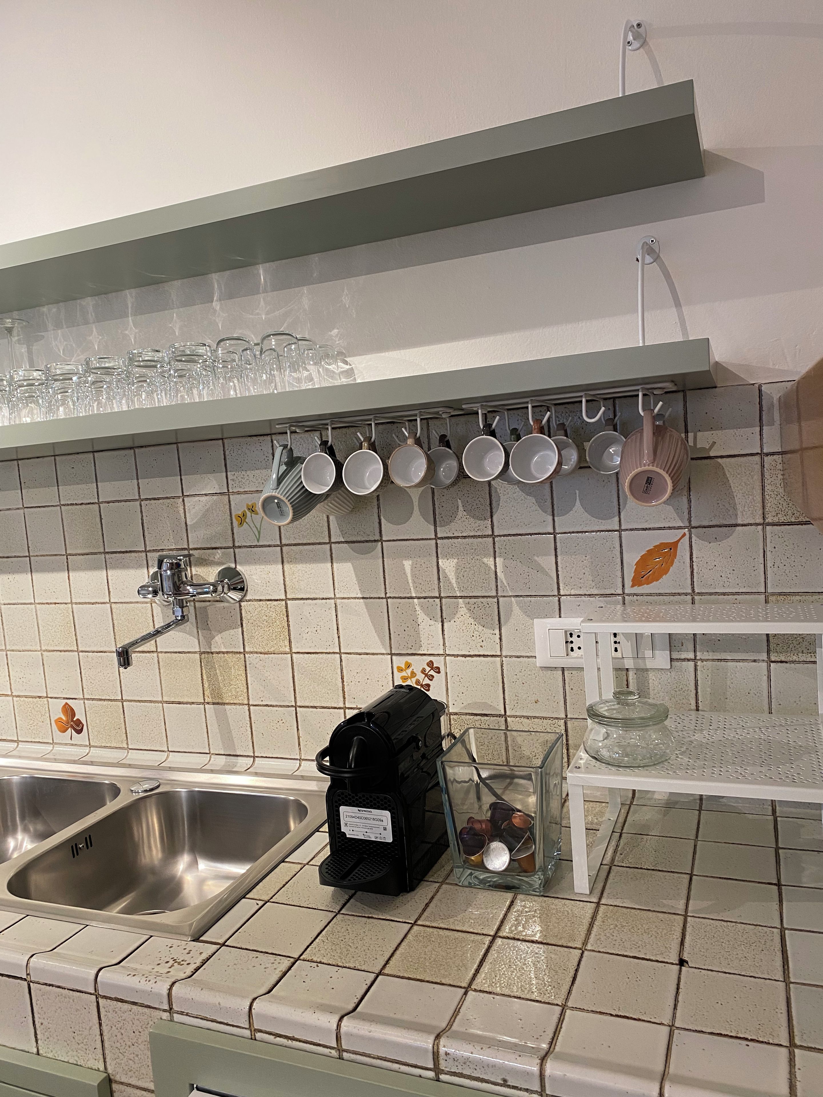 A kitchen with two stainless steel sinks, a coffee maker, a glass jar, and a shelf with various kitchenware.