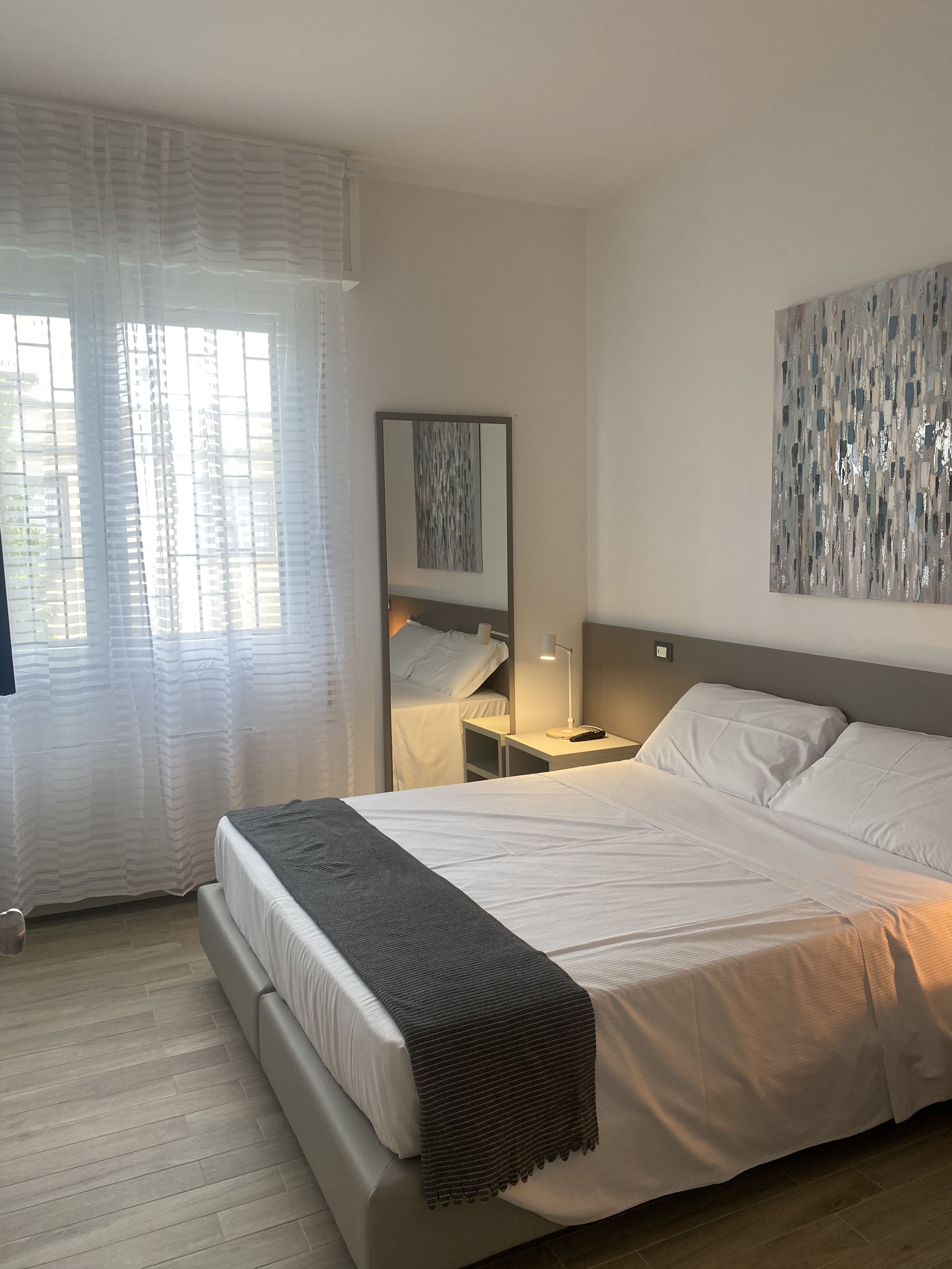 A hotel room with a bed, a bedside table with a lamp, a mirror, and a window with curtains.