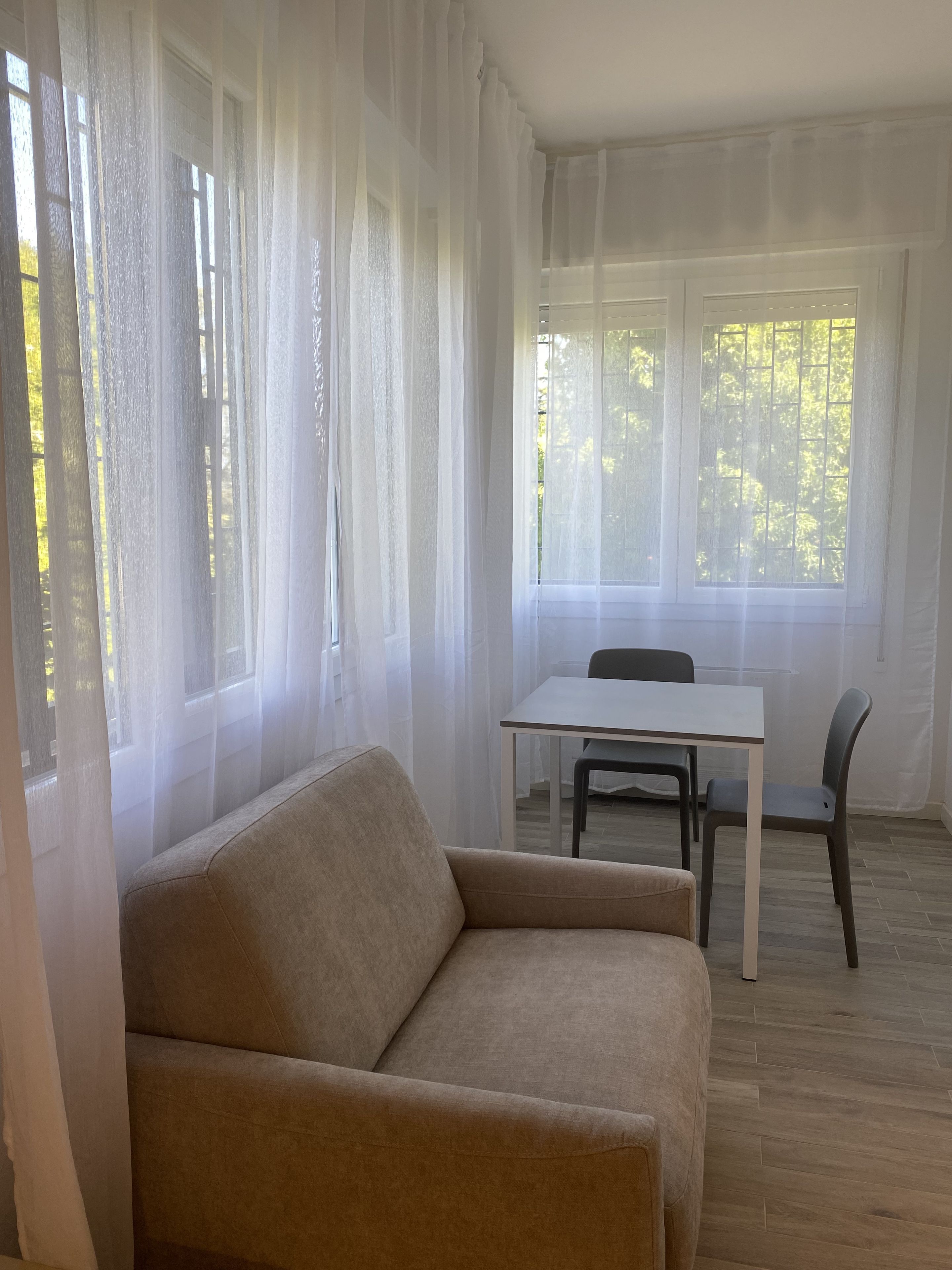 Una habitación con un sofá color beige, una mesa blanca y dos sillas. Hay ventanas amplias con cortinas de gasa.