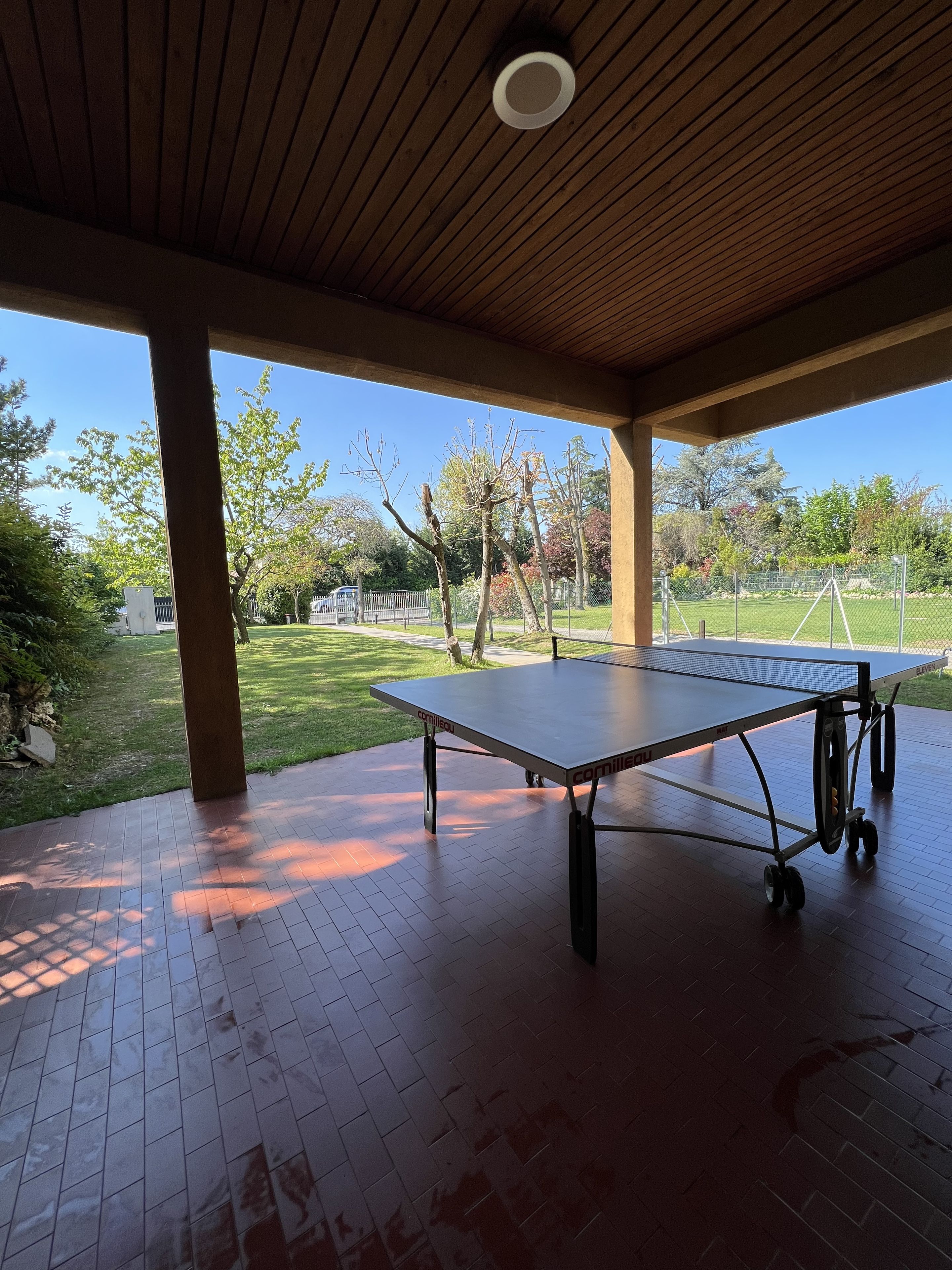 An outdoor ping pong table under a wooden pavilion with a clear view of a grassy area and trees.