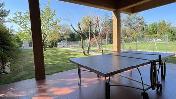 Ein Outdoor-Tischtennis-Tisch unter einer hölzernen Laube mit freiem Blick auf eine Rasenfläche und Bäume.
