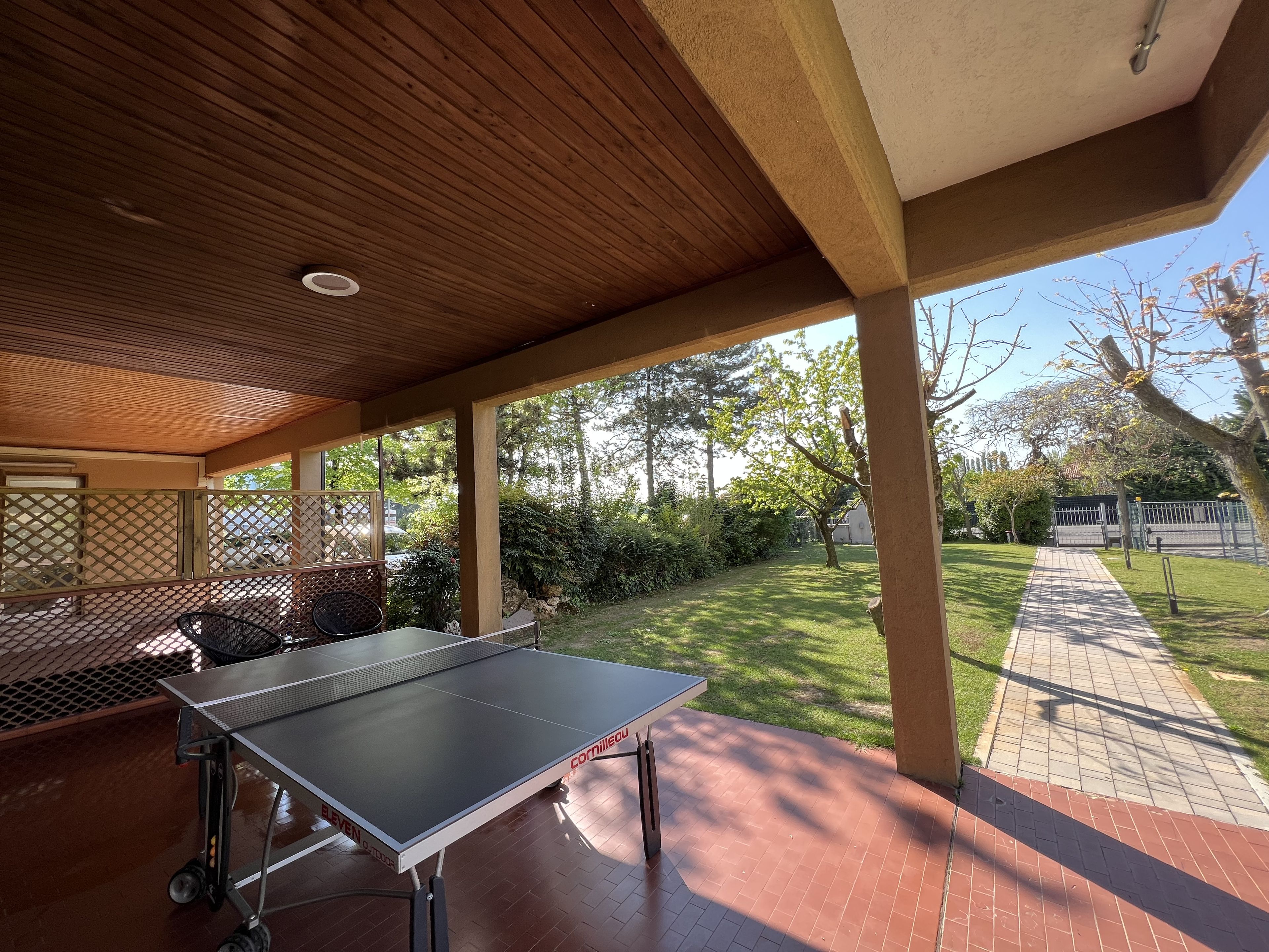 An outdoor ping pong table under a wooden pergola with a view of a well-maintained lawn and trees.