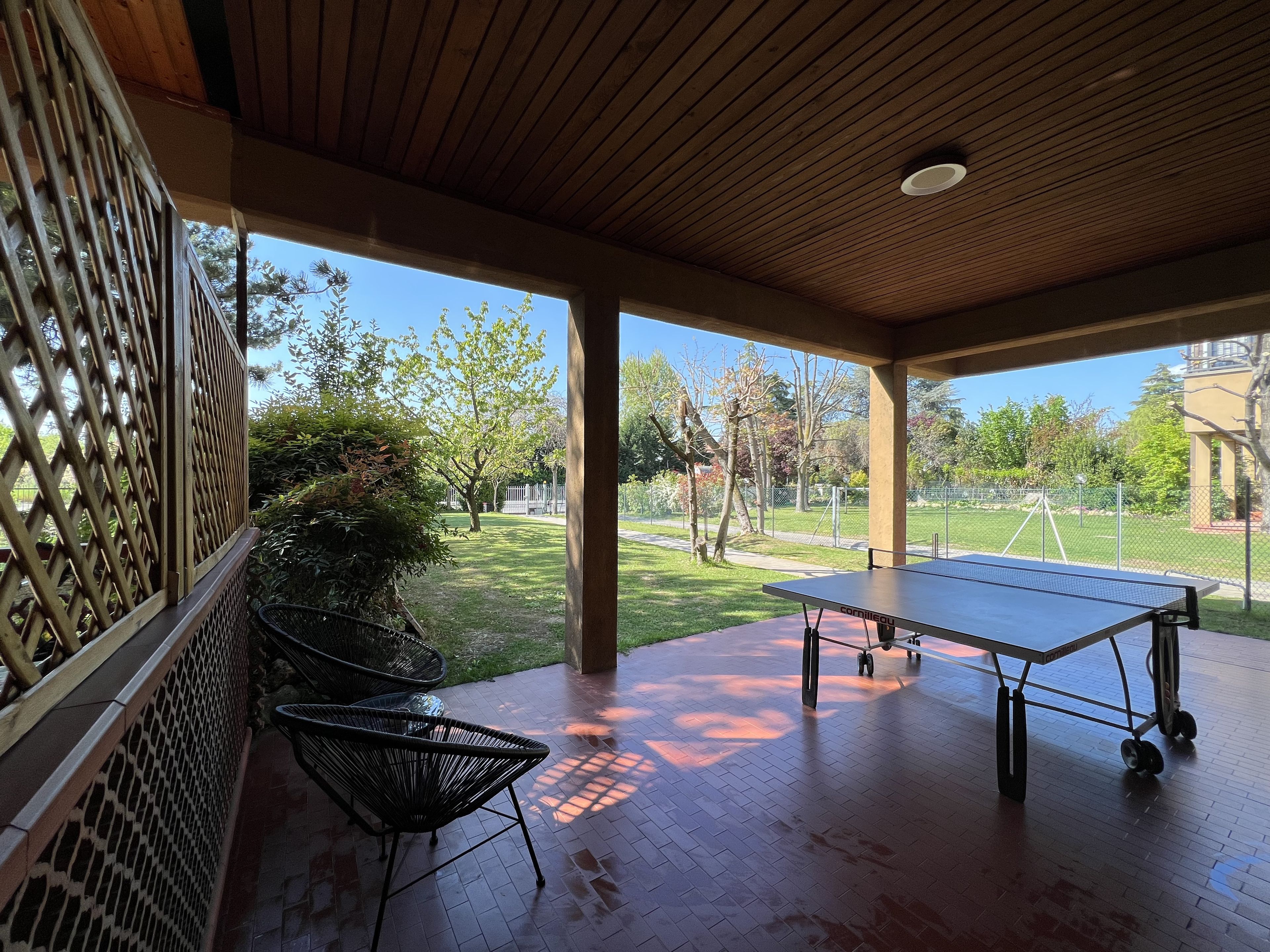 Un espace extérieur abrité comprenant une table de ping-pong, des chaises et une vue sur un parc avec des arbres et une clôture.