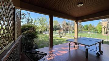 Un espace extérieur abrité comprenant une table de ping-pong, des chaises et une vue sur un parc avec des arbres et une clôture.