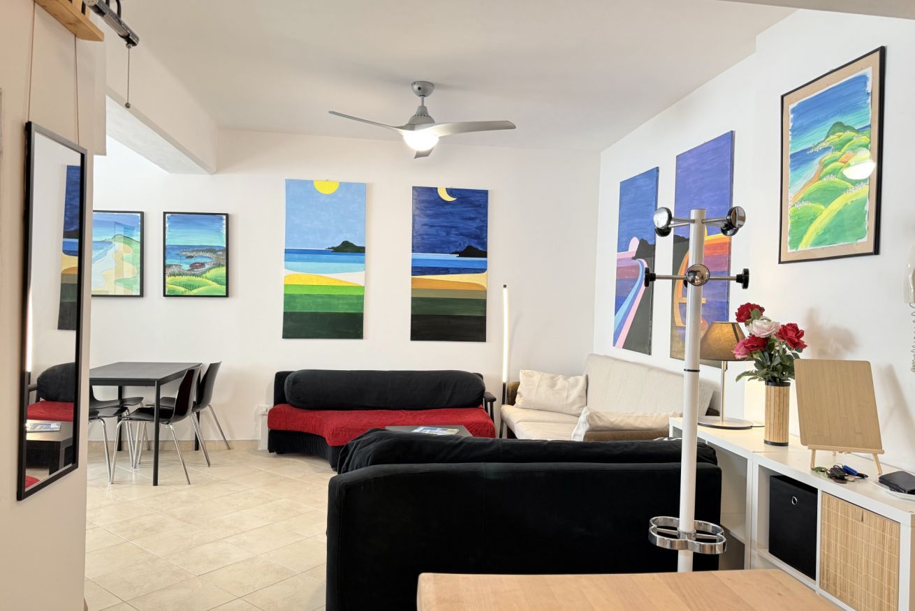 A modern living room with a black sofa, a dining table, and chairs. There are paintings on the walls and a ceiling fan.