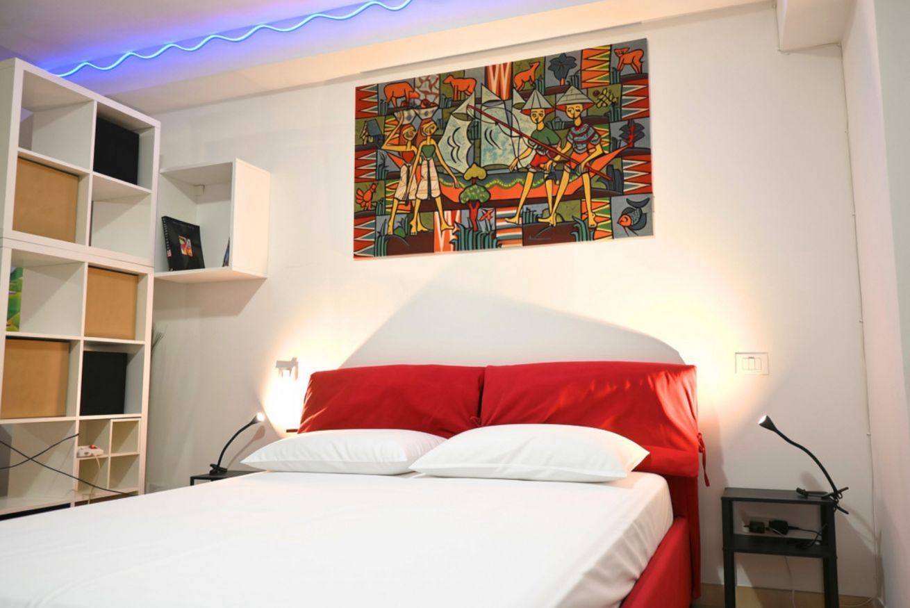 Une chambre à coucher avec un lit, une tête de lit blanche, une touche de couleur rouge, un tableau accroché au mur et une étagère.