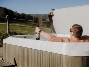 A person relaxing in a hot tub outdoors with a bottle of beer, surrounded by a wooden deck and a view of a grassy field and hills. - One-bedroom Cabin (Sankt Georgen am Kreischberg)