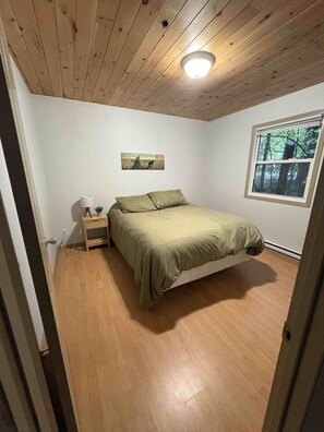 Un dormitori amb un llit, una tauleta de nit, una làmpada, un quadre a la paret i una finestra amb vista a arbres.