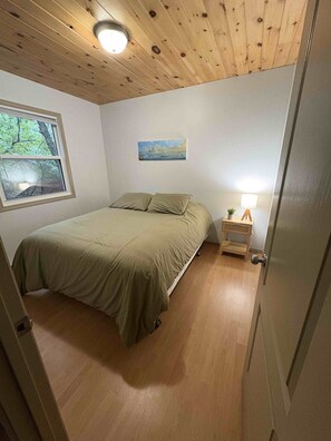 Quarto com cama, mesa de cabeceira com abajur, um quadro na parede e janela com vista para árvores.