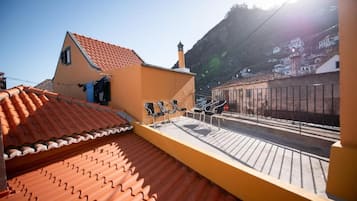 A rooftop terrace with a tiled roof, a small building with a chimney, and a view of a street and hills in the background.