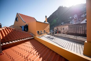 Una terraza en la azotea con tejado de baldosas, un edificio pequeño con chimenea y vistas a una calle y colinas al fondo.