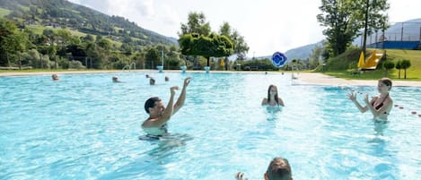 A swimming pool with multiple people in the water, surrounded by a landscape with trees and hills.