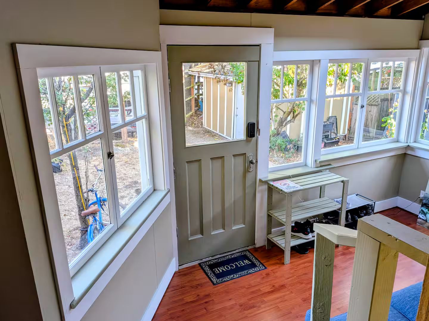 A room with a door, windows, a welcome mat, and a small table with shelves.