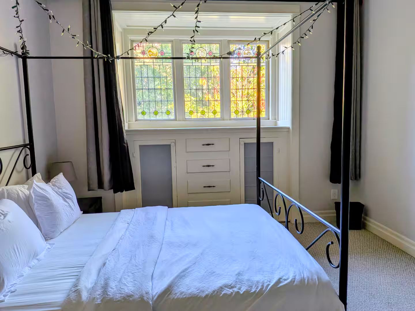 Un lit à baldaquin, une fenêtre ornée de vitraux et une commode blanche à tiroirs.