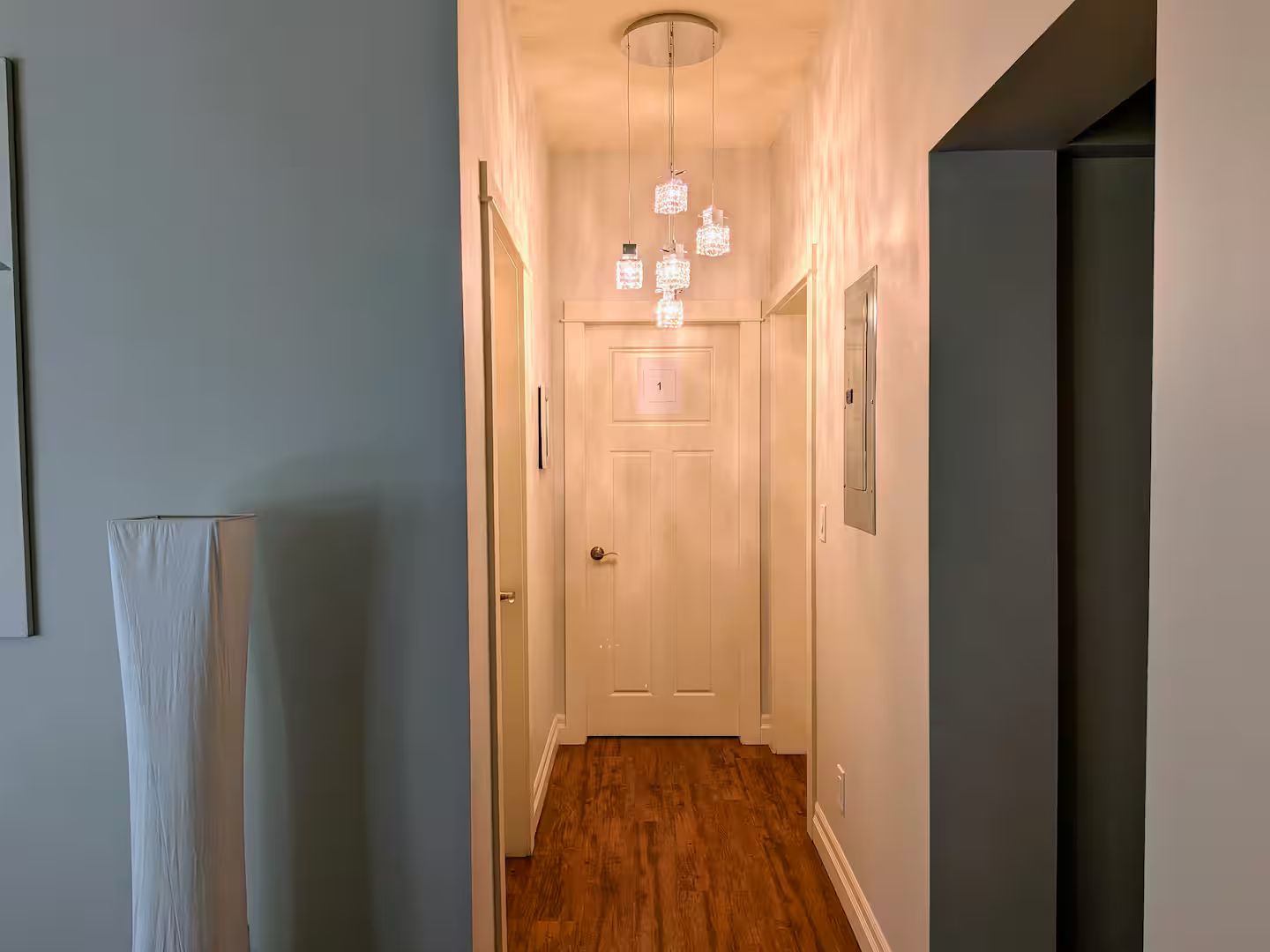 A hallway with a wooden floor, a white door, and a chandelier with multiple lights.