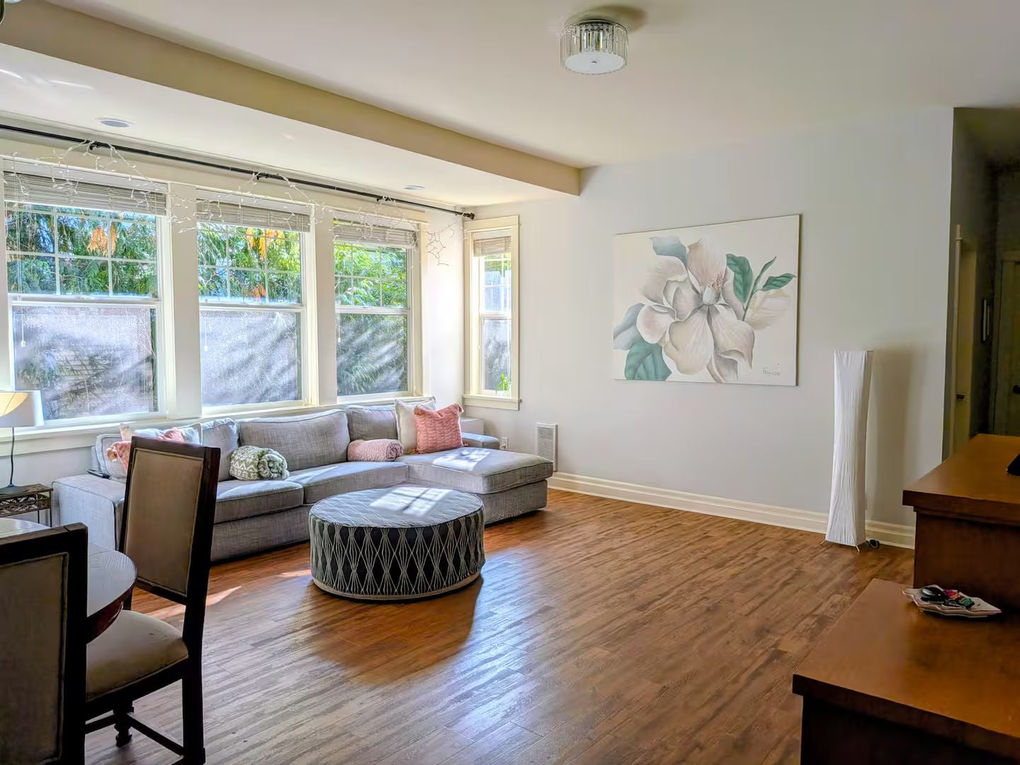 A spacious living room with a large window, a round ottoman, a sofa, and a painting on the wall.