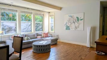 A spacious living room with a large window, a round ottoman, a sofa, and a painting on the wall.