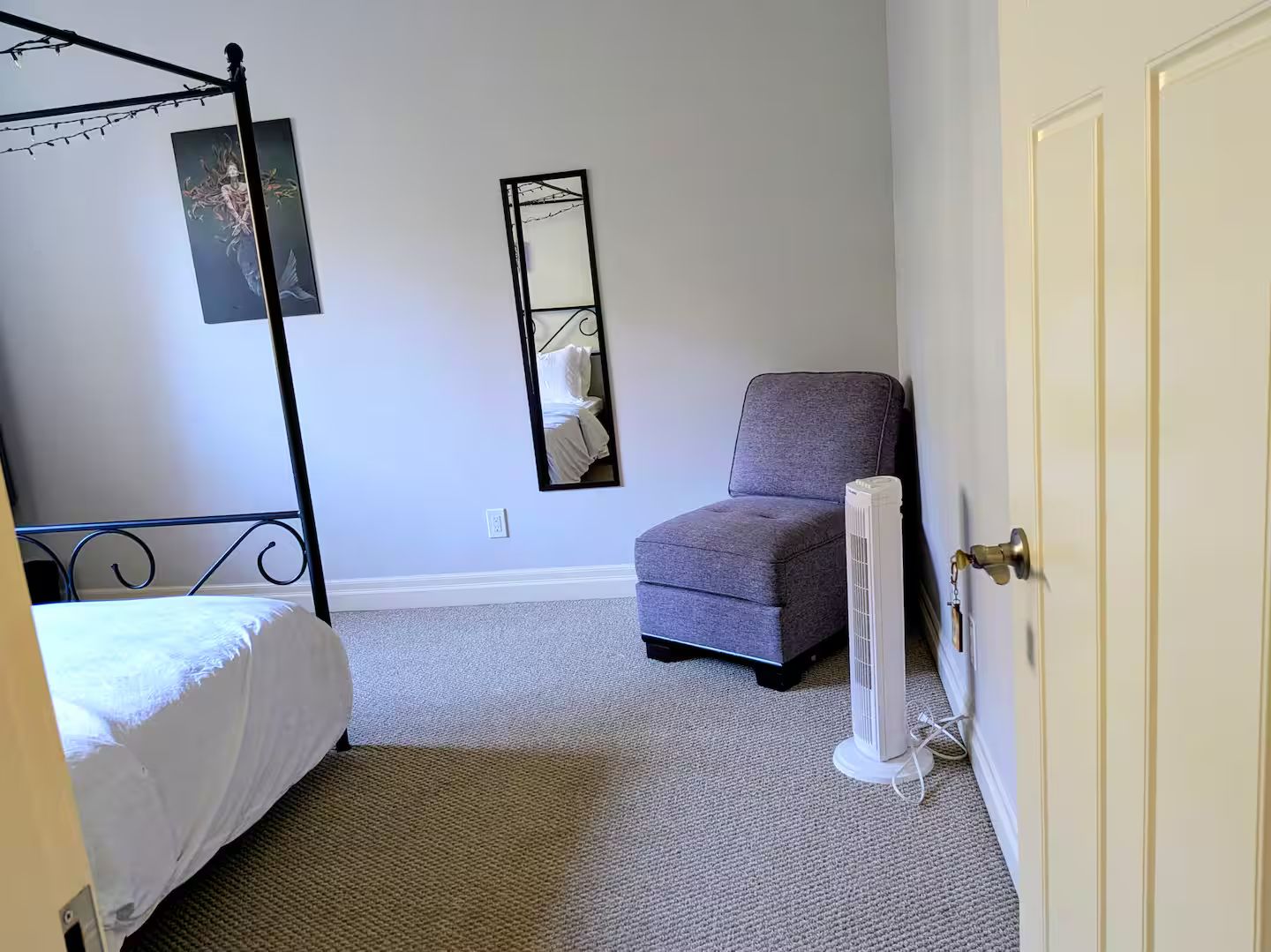 A bedroom with a four-poster bed, a chair, a standing heater, and a mirror on the wall.