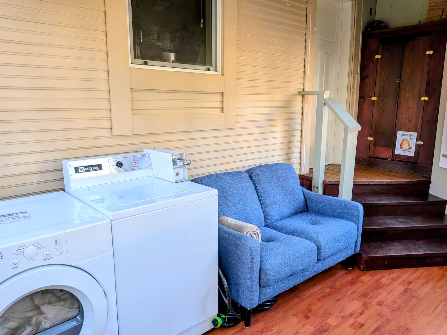 A laundry area with a front-loading washer and dryer, a blue sofa, and a wooden staircase leading to a door with a 'Shoes Off' sign.