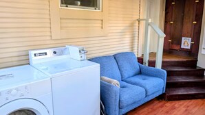 A laundry area with a front-loading washer and dryer, a blue sofa, and a wooden staircase leading to a door with a 'Shoes Off' sign. - Comfortable private room near downtown Victoria (Victoria)