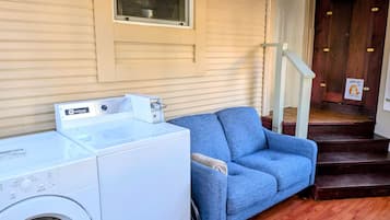 A laundry area with a front-loading washer and dryer, a blue sofa, and a wooden staircase leading to a door with a 'Shoes Off' sign.