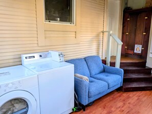 A laundry area with a front-loading washer and dryer, a blue sofa, and a wooden staircase leading to a door with a 'Shoes Off' sign. - Comfortable private room near downtown Victoria (Victoria)